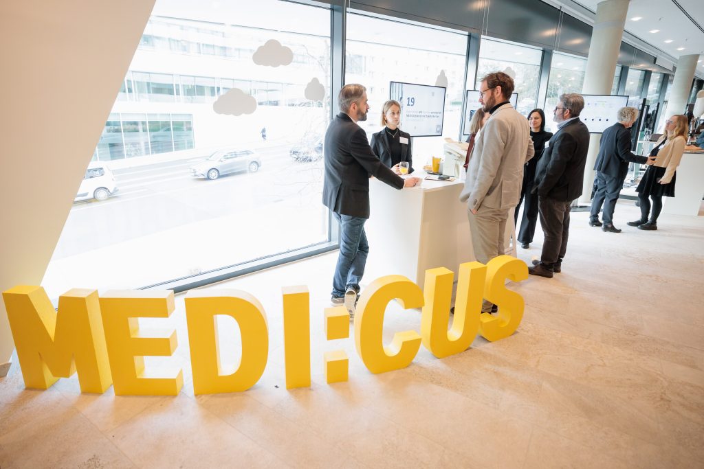 Mehrere Personen stehen während der Jahresveranstaltung im Forum Gesundheitsstandort vor großen Fenstern und unterhalten sich. Im Vordergrund ist ein großes gelbes MEDI:CUS-Schild zu sehen.