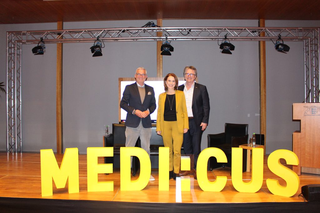 Beim Event Daten retten Leben im März 2025 stehen Thomas Strobl, Petra Olschowski und Manfred Lucha auf der Bühne. Im Vordergrund sind große gelbe Buchstaben zu sehen mit dem Projektnamen MEDI:CUS.