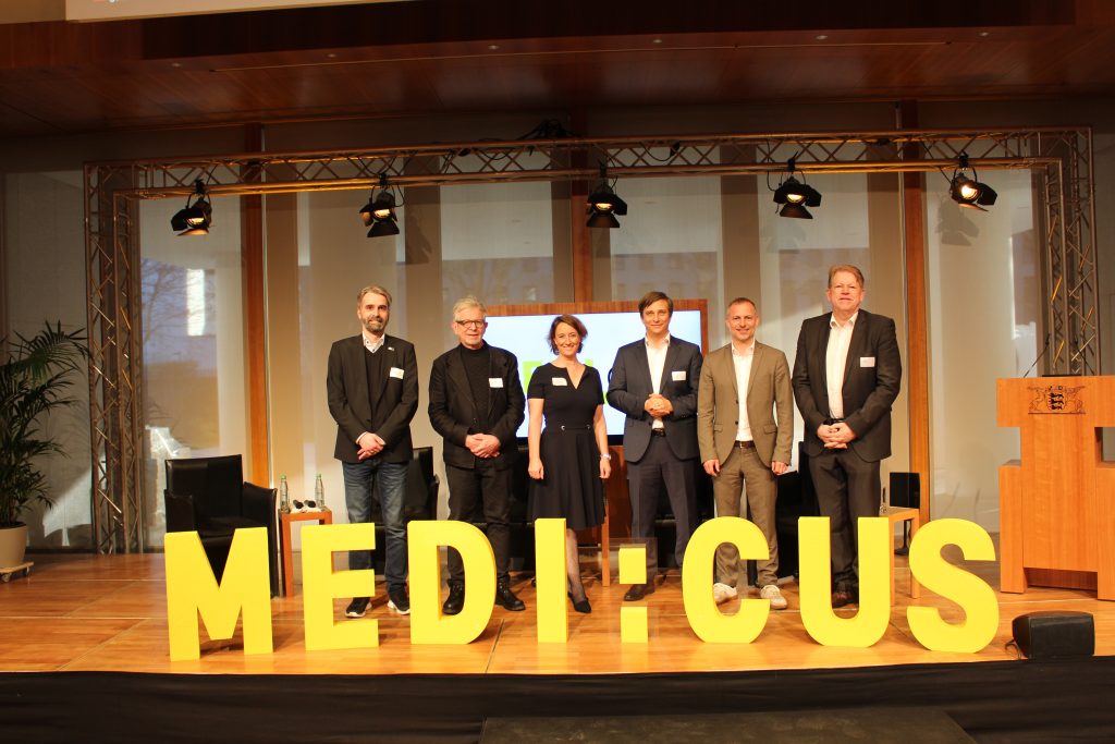 Beim Event Daten retten Leben im März 2025 stehen die Projektverantwortlichen Alexander Beckee, Prof. Dr. Jörg Martin, Eva Schulz, Prof. Dr. Dr. Martin Holderried, Matthias Pröfrock und Klaus Mühleck auf der Bühne. Im Vordergrund sind große gelbe Buchstaben zu sehen mit dem Projektnamen MEDI:CUS.
