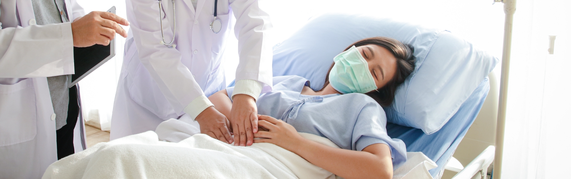 Ärztliche Fachperson untersucht im Bett liegende Person am Bauch, weitere ärztliche Person steht daneben mit Tablet