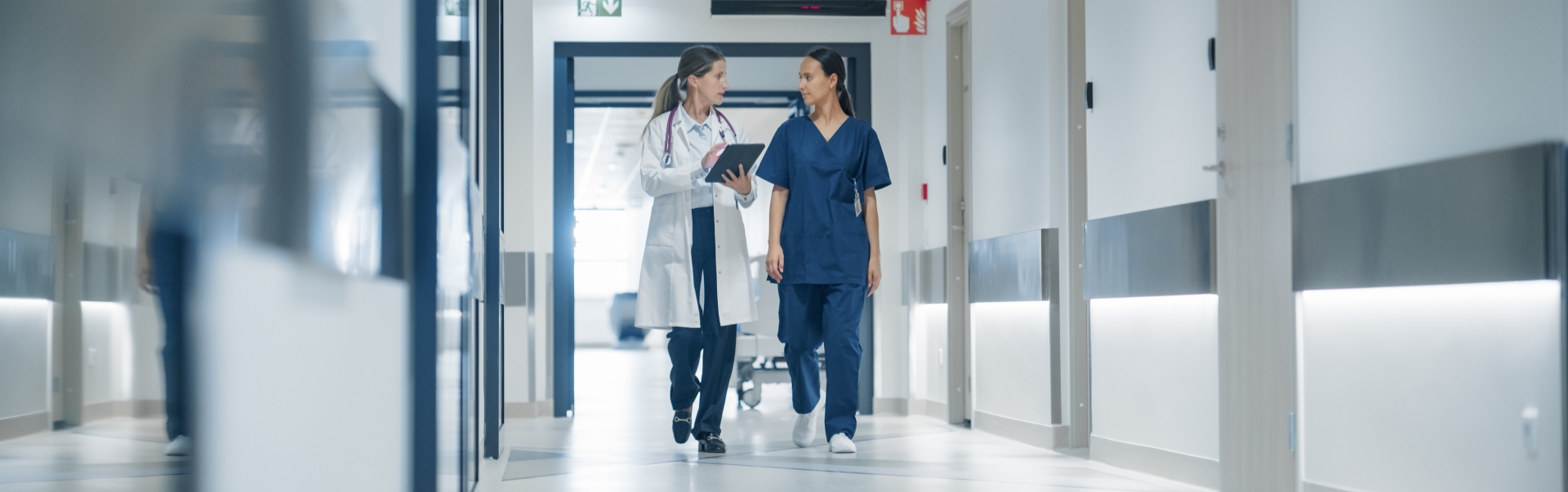Zwei Medizinerinnen, eine im weißen Kittel, eine andere in blauer Kleidung, sprechen über MEDI:CUS-Lösungen, während sie durch einen hellen, modernen Krankenhausflur gehen.