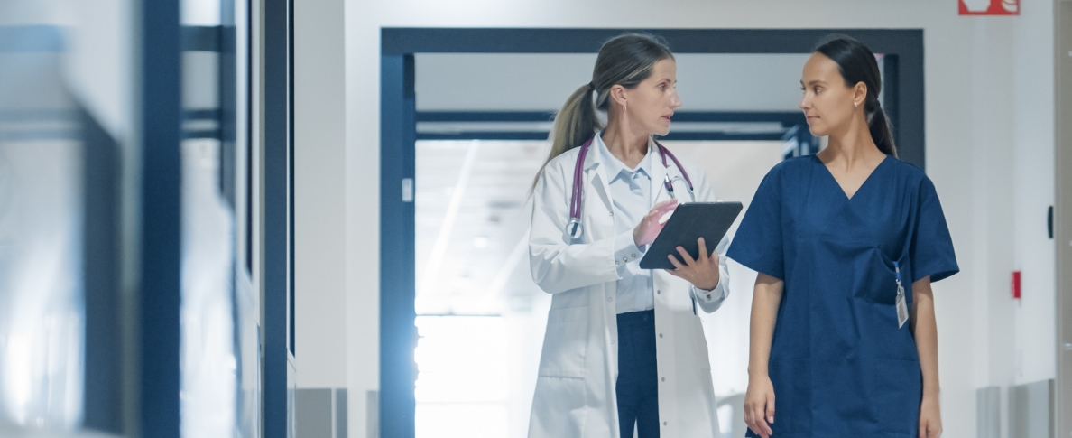 Zwei Medizinerinnen, eine im weißen Kittel, eine andere in blauer Kleidung, sprechen über MEDI:CUS-Lösungen, während sie durch einen hellen, modernen Krankenhausflur gehen.